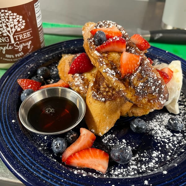 Golden french toast with strawberries, blueberries, powdered sugar, whipped cream, and maple syrup on a blue plate.