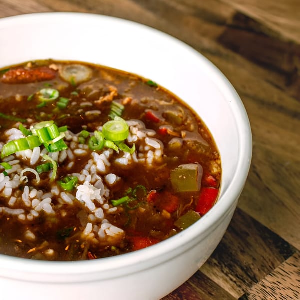 Close-up of a bowl of dark Cajun gumbo with white rice, green onions, and vegetables.