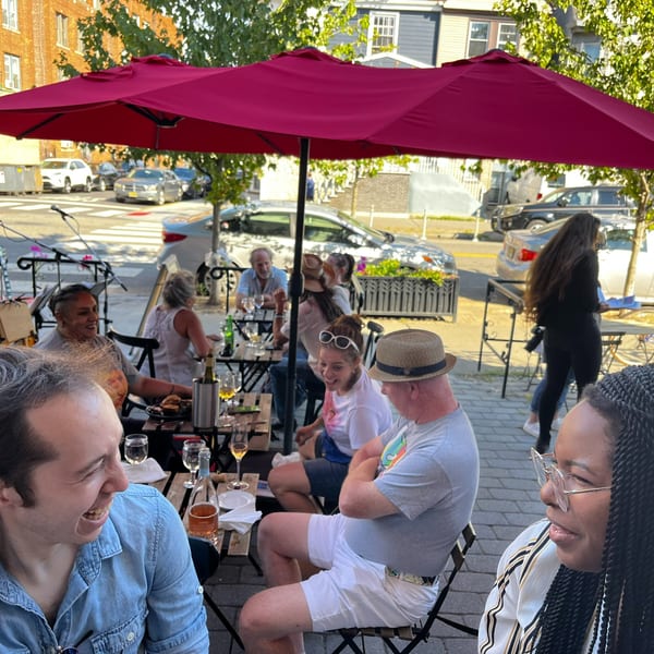 People enjoying drinks and conversation at an outdoor restaurant patio under red umbrellas.