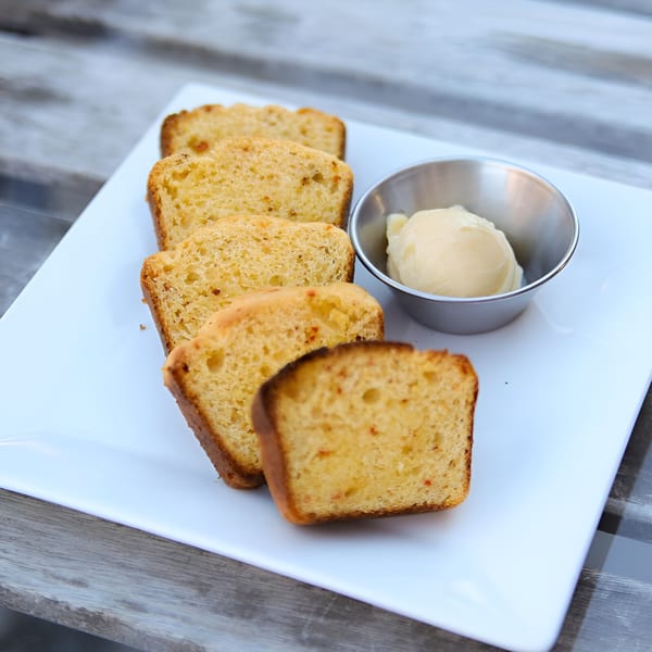 Slices of golden cornbread and a small metal cup of butter on a white rectangular plate.